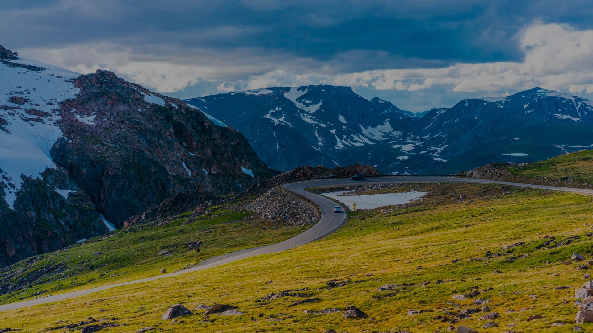 A wide landscape view of a scenic highway cutting through snow-peaked mountains and green meadows